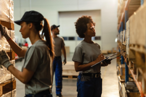 A South African woman inspects boxes in a warehouse with a clipboard.
